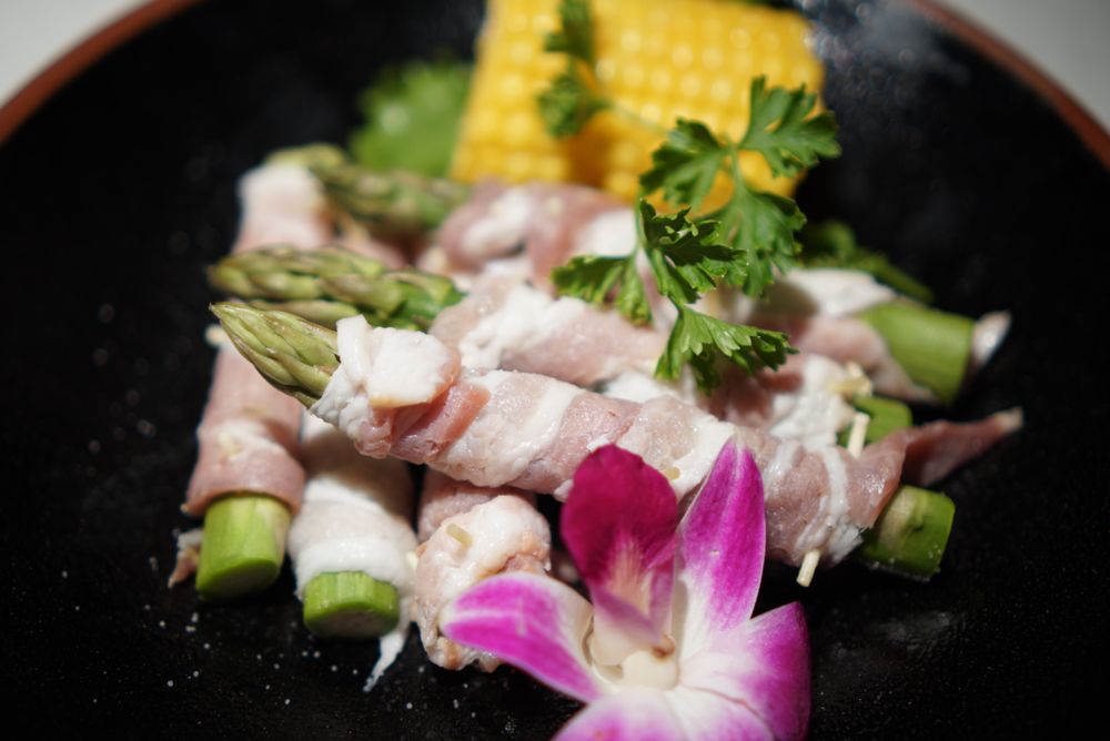 Bacon-wrapped asparagus rolls on a black plate with orchid garnish and corn